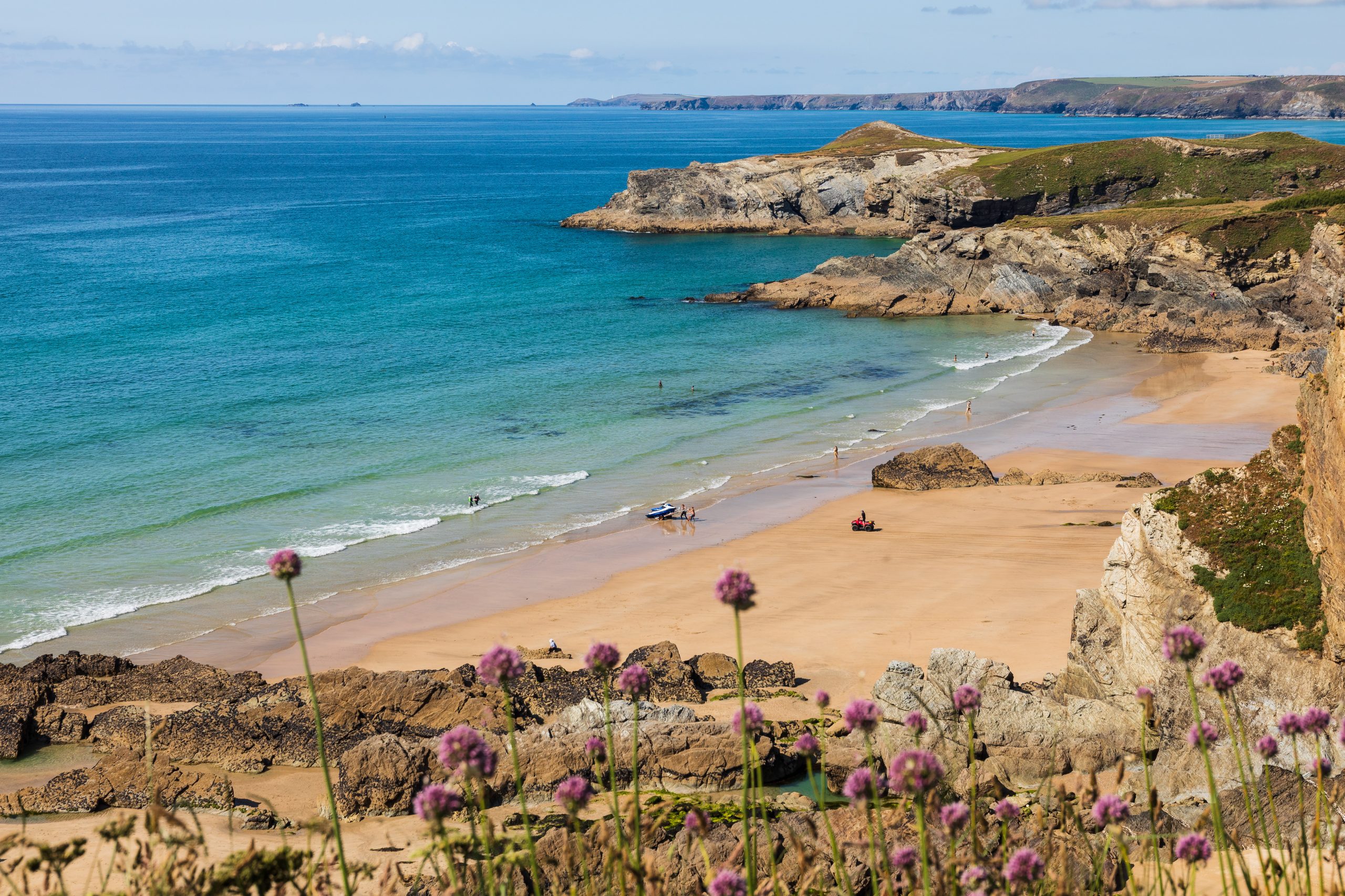 Clear turquoise waves gently roll onto the light golden sands of Lusty Glaze Beach with Porth Island and Newquay Bay beyond.